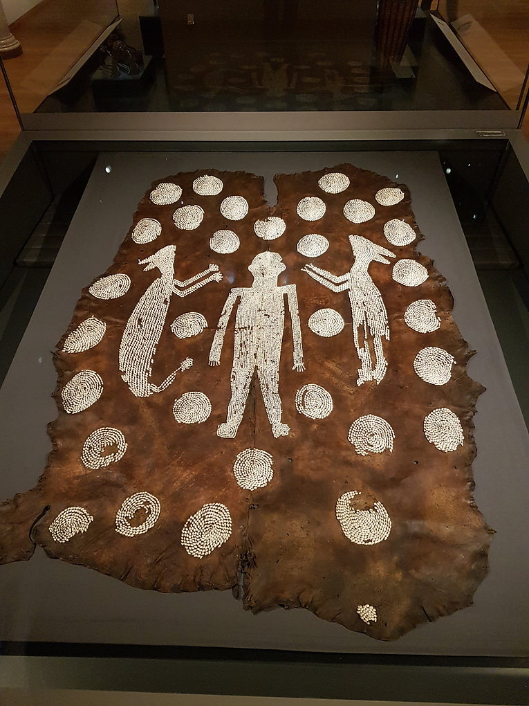 Photograph showing a mantle made of leather or skin and decorated with embroidered shells in a glass case.